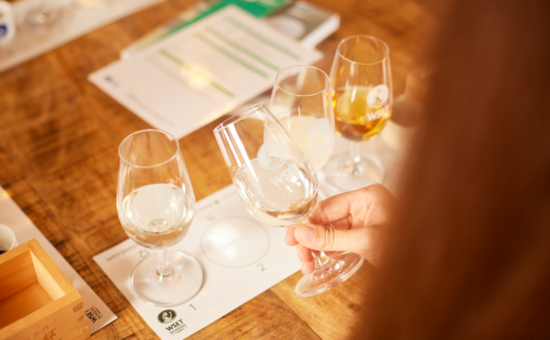 A student holding a wine glass filled with sake above a white mat on a wooden table