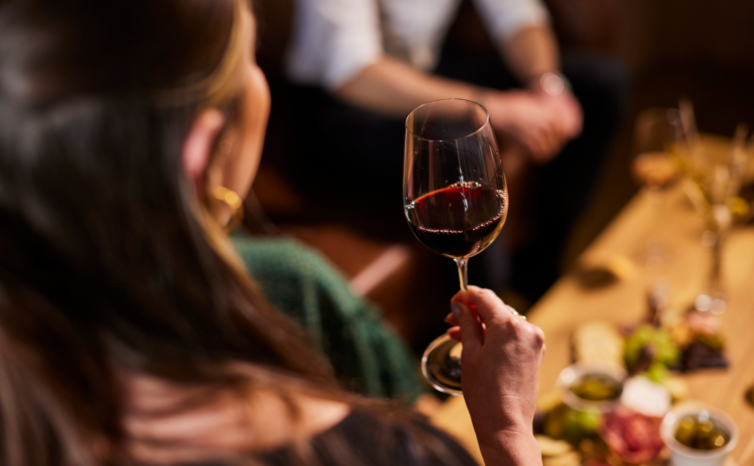 A person holding a glass of red wine, with cheese and charcuterie snacks in the background.