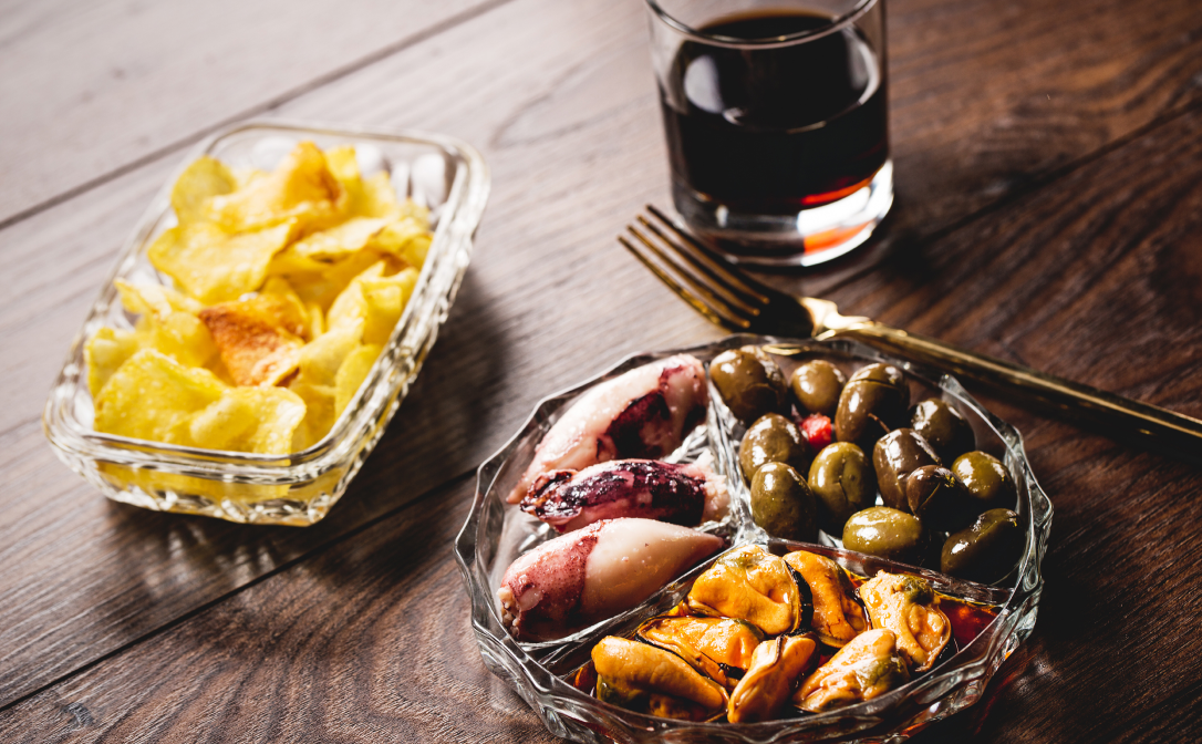 A glass of vermouth on a wooden table, with snacks like crisps and olives