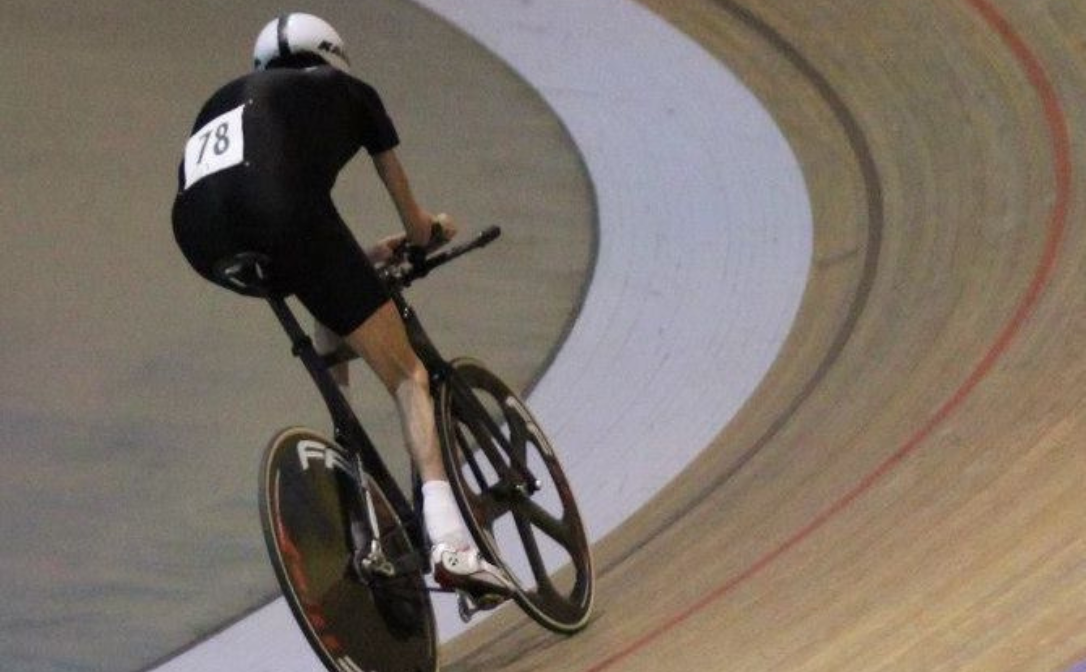 A person clothed in a tight black cycling outfit, racing on a bike track