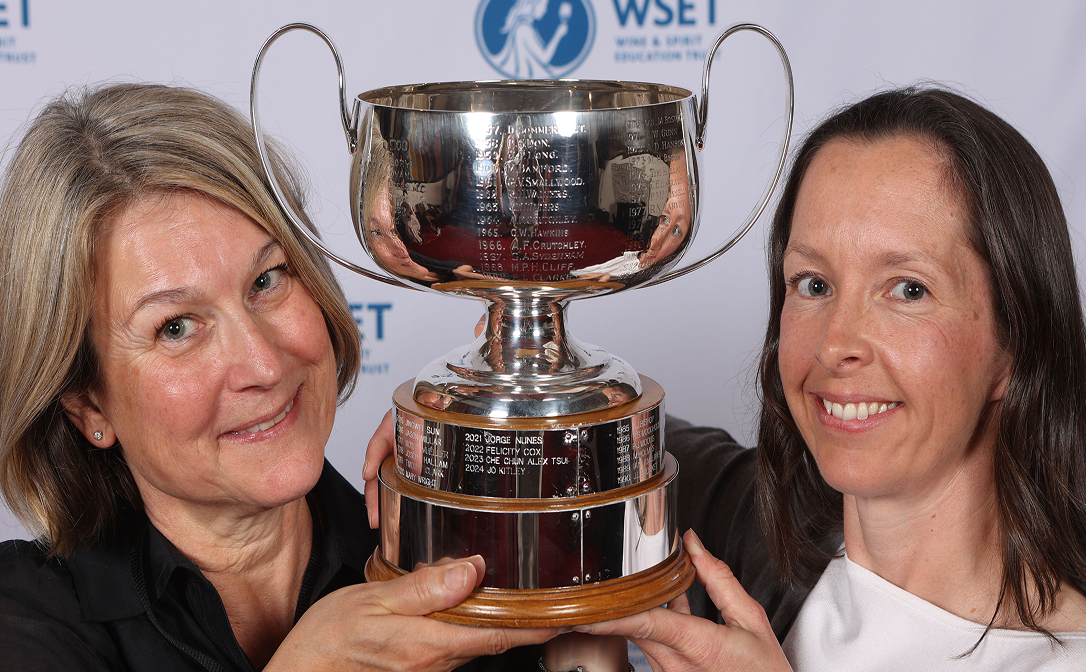 Two women in front of a white and blue banner, holding a large silver trophy