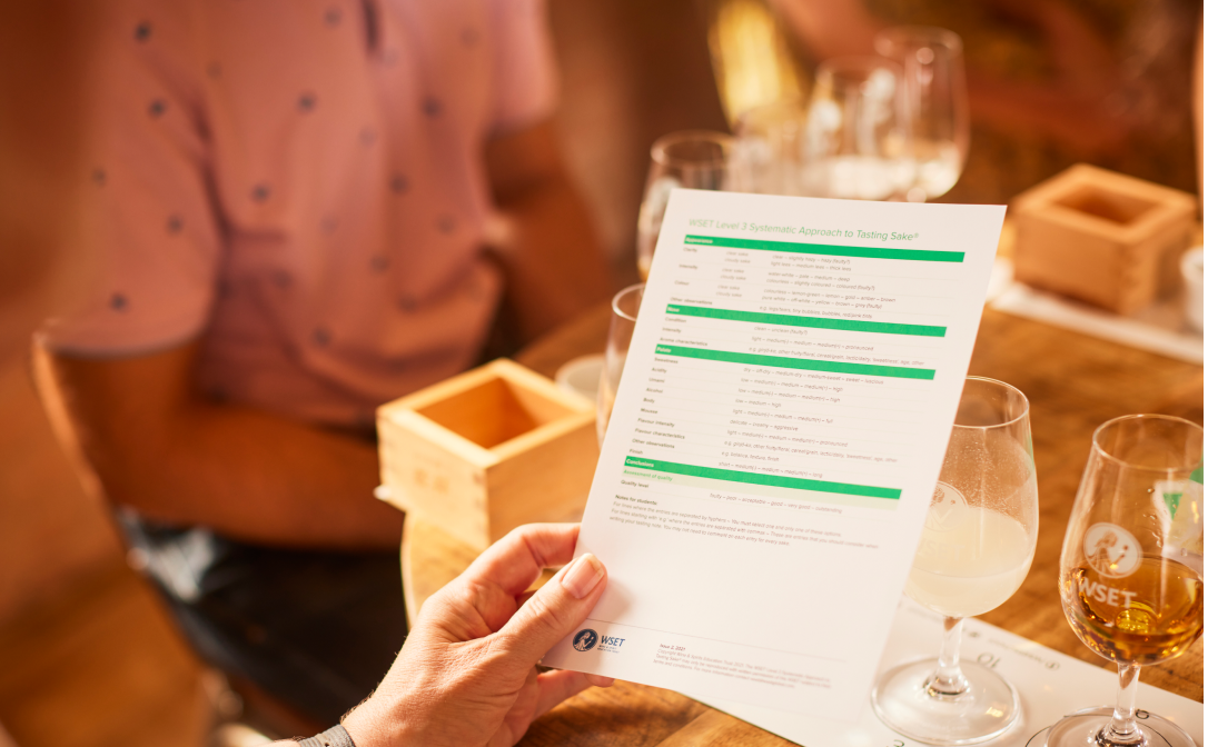 A white and green document held in someone's hand, with small glasses filled with different colour liquids in the background